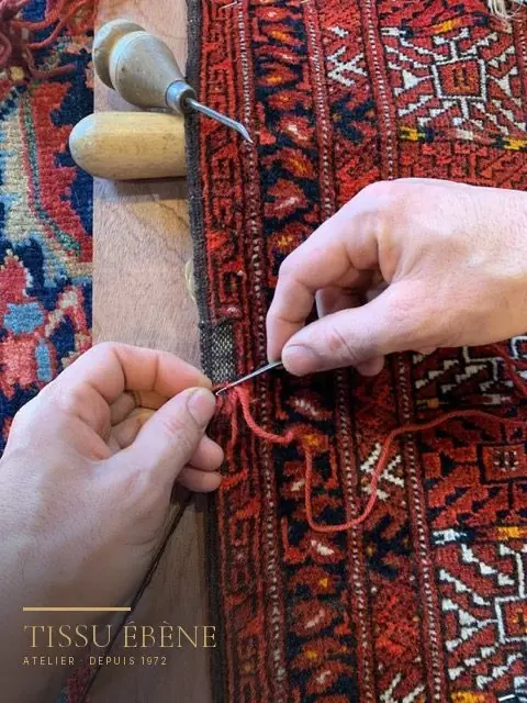 Mains cousant au fil rouge la lisière d’un tapis en laine à motifs rouges, outils en bois à côté